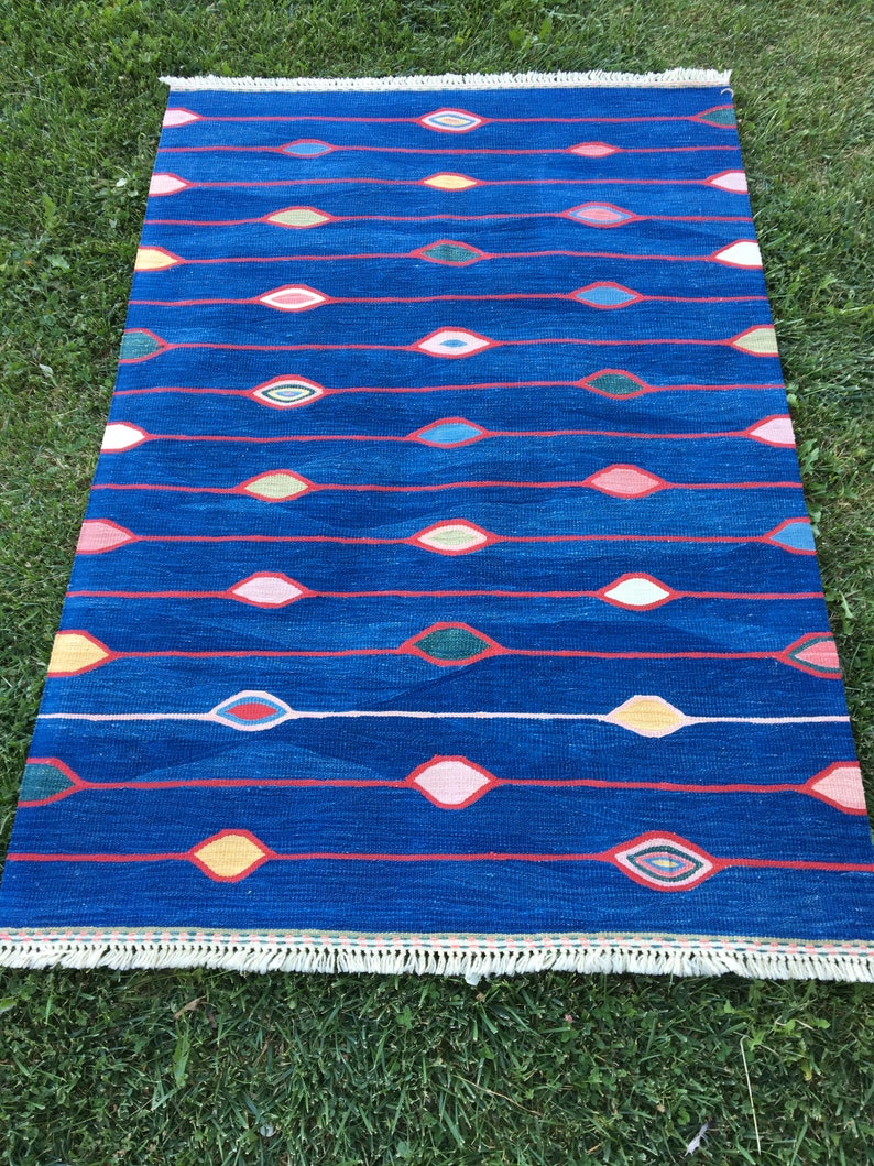 Puede incluir: Una alfombra azul con un patr&oacute;n geom&eacute;trico de &oacute;valos rojos, rosas, amarillos y verdes. La alfombra tiene un borde de flecos blancos.
