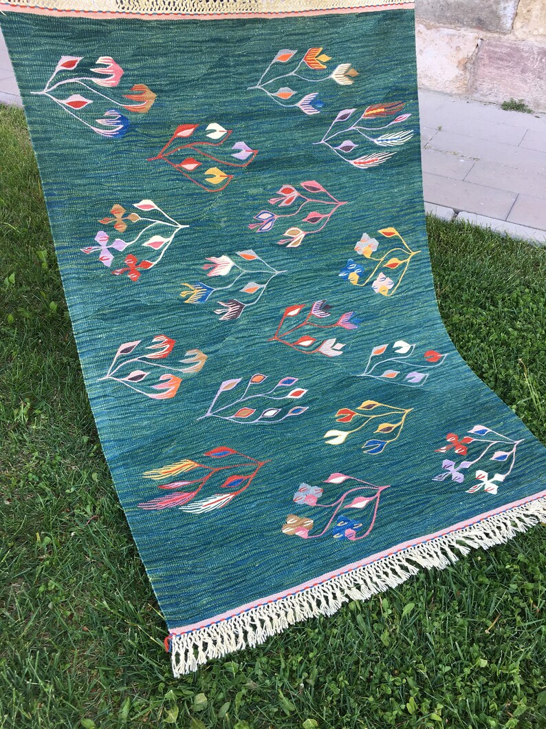 Puede incluir: Una alfombra azul verdosa con borde de flecos presenta un patr&oacute;n repetitivo de dise&ntilde;os florales estilizados en tonos de rosa, amarillo, verde, azul y naranja.