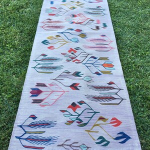 May include: A long, rectangular rug with a white background and a colorful geometric pattern of stylized flowers and leaves. The rug has a fringe border.