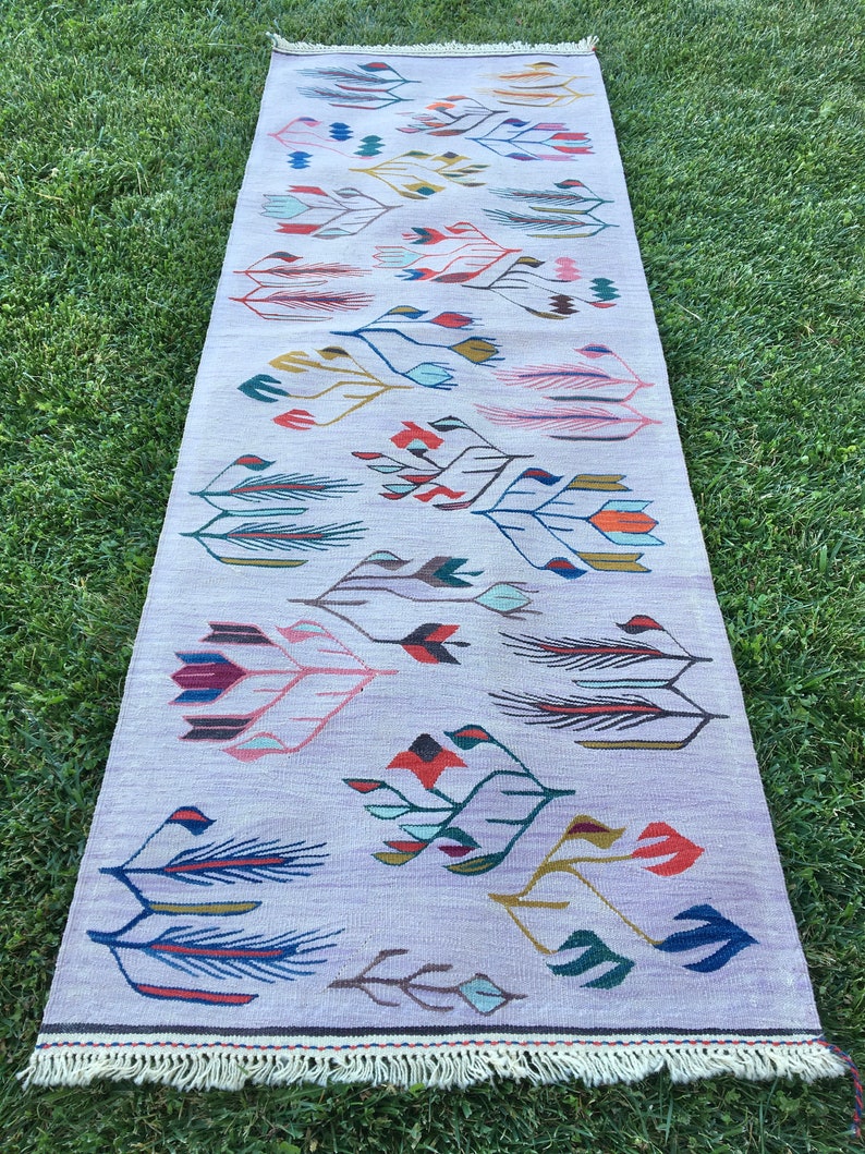 May include: A long, rectangular rug with a white background and a colorful geometric pattern of stylized flowers and leaves. The rug has a fringe border.