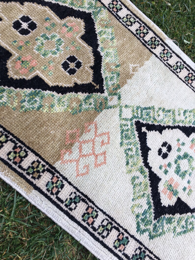 May include: A vintage rug with a geometric pattern in shades of green, black, and pink. The rug has a white background and a black border.