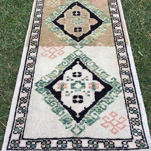 May include: A white and beige rug with a geometric pattern in black, green, and pink. The rug has a fringed border.