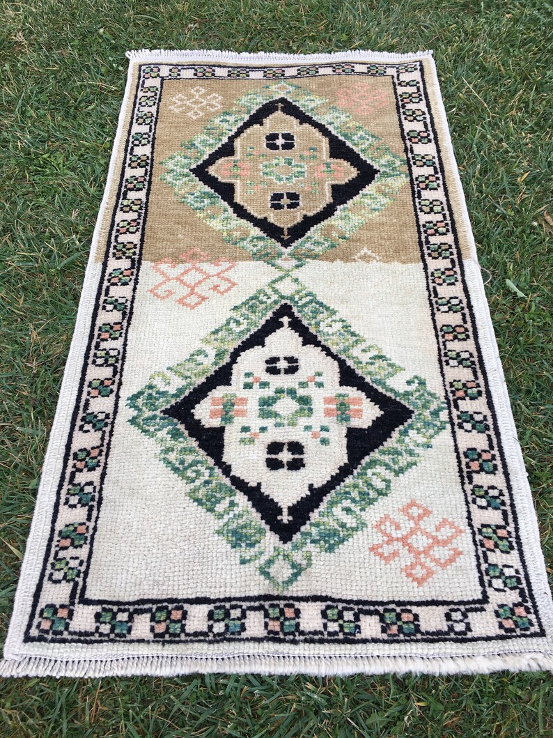 May include: A white and beige rug with a geometric pattern in black, green, and pink. The rug has a fringed border.
