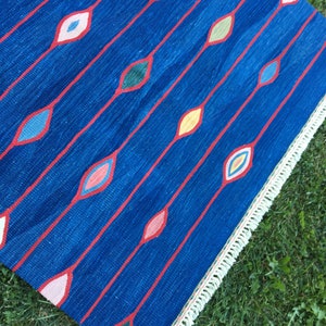 Puede incluir: Una alfombra azul con un patr&oacute;n geom&eacute;trico de diamantes de colores. La alfombra tiene un borde de flecos blancos.
