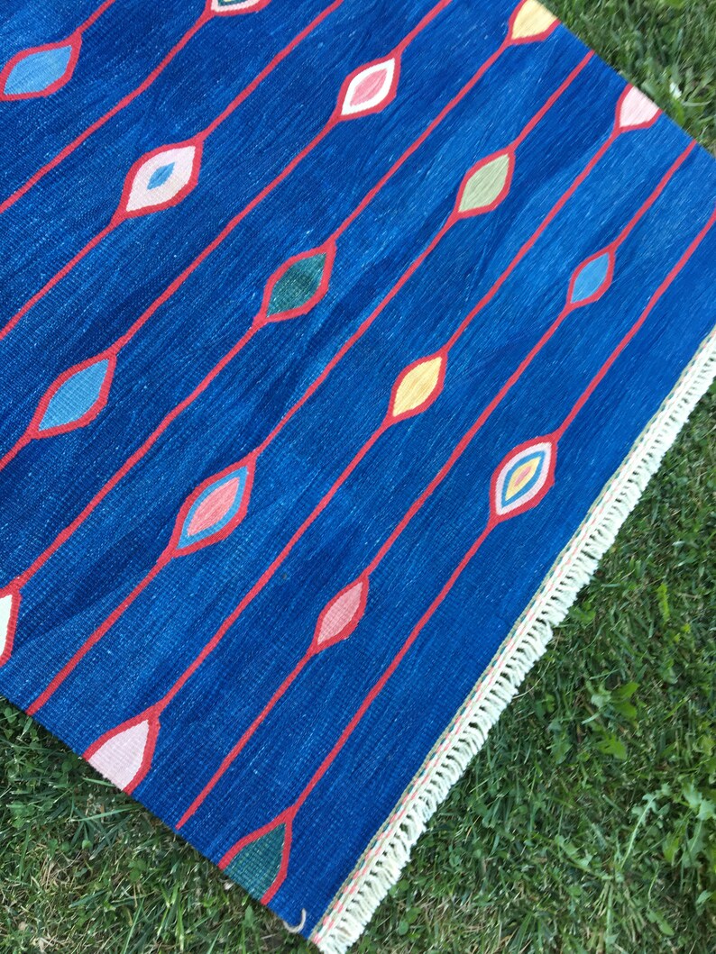 Puede incluir: Una alfombra azul con un patr&oacute;n geom&eacute;trico de diamantes de colores. La alfombra tiene un borde de flecos blancos.