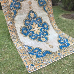 May include: A rectangular, vintage-style rug with a central floral medallion in shades of blue and gold. The rug has a cream-colored background and a detailed border. The rug is displayed on a green lawn.