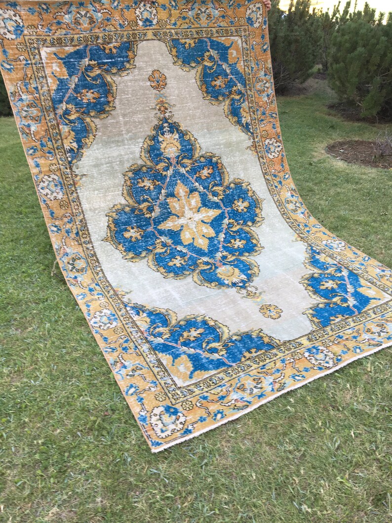 May include: A rectangular, vintage-style rug with a central floral medallion in shades of blue and gold. The rug has a cream-colored background and a detailed border. The rug is displayed on a green lawn.