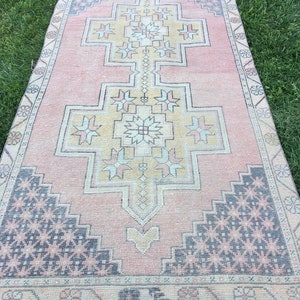 May include: A rectangular rug with a central geometric design in shades of pink, beige, and yellow. The rug features a border with a repeating pattern and dark blue accents. The rug is placed on a green grassy surface.