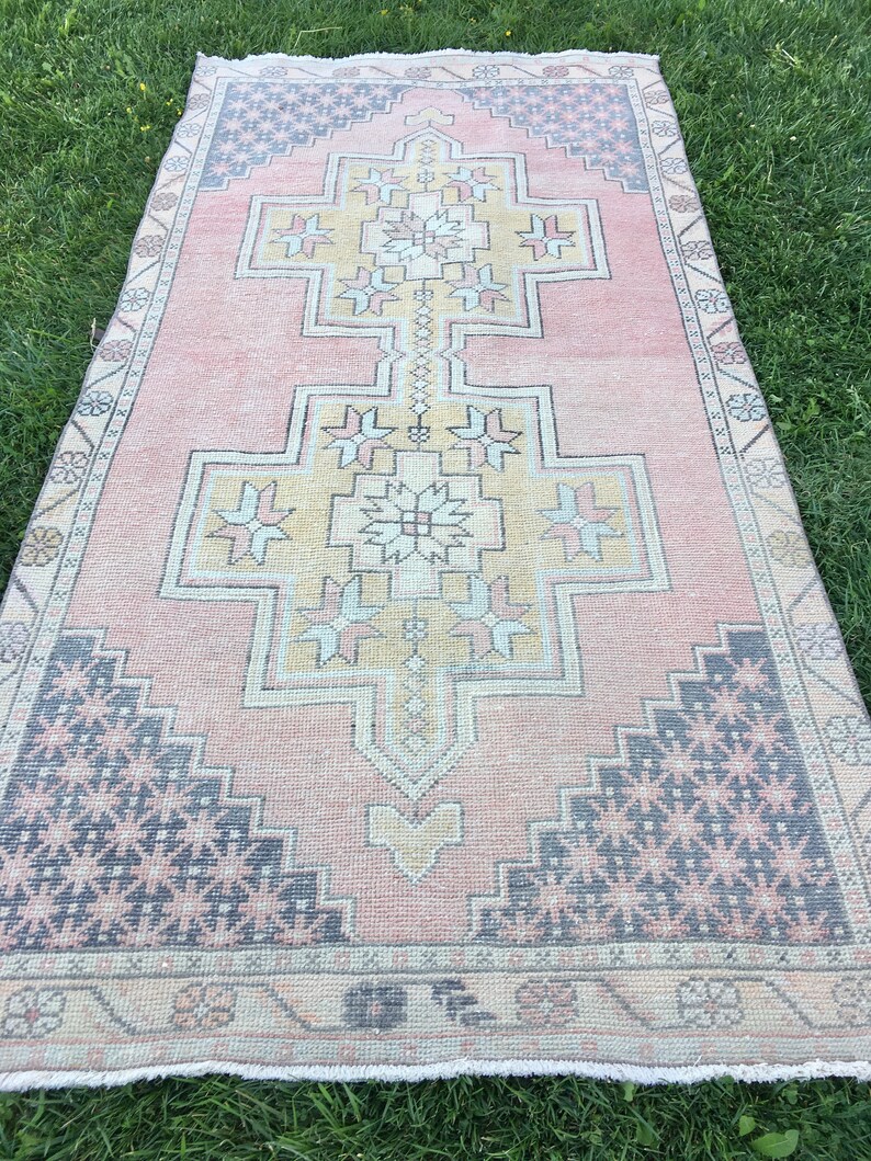 May include: A rectangular rug with a central geometric design in shades of pink, beige, and yellow. The rug features a border with a repeating pattern and dark blue accents. The rug is placed on a green grassy surface.