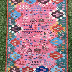 May include: A colourful rug with a pink background and a floral pattern. The rug has a green border and white fringe.