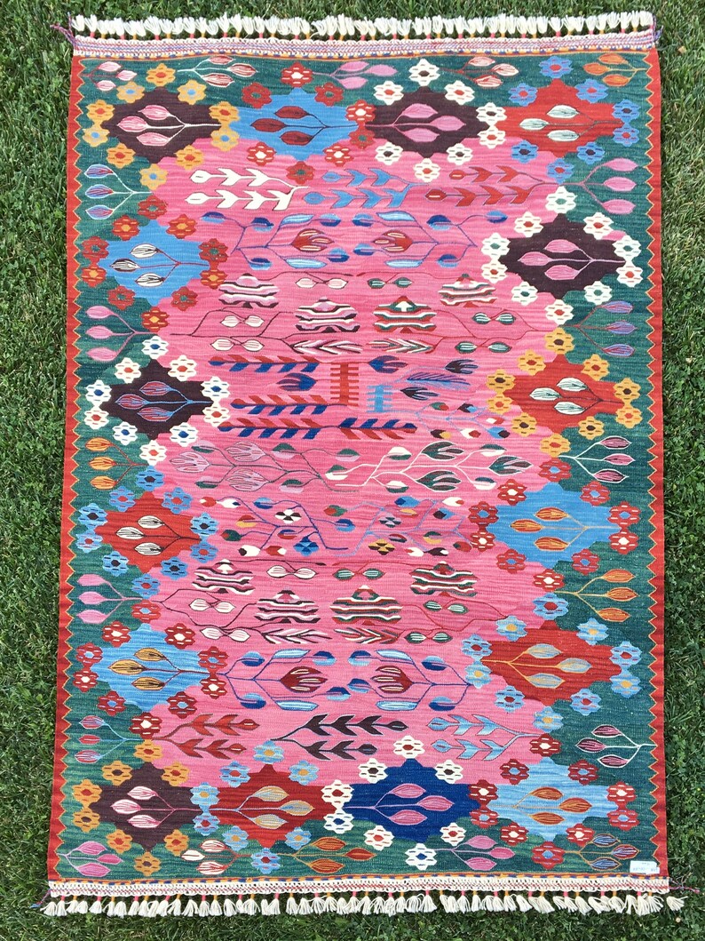 May include: A colourful rug with a pink background and a floral pattern. The rug has a green border and white fringe.
