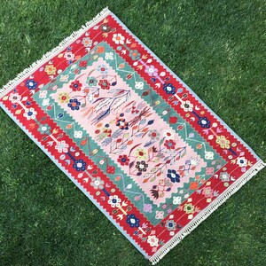 May include: A rectangular rug with a floral design. The rug features a pink central field with colorful flowers and a green border. The outer border is red with white fringe on the edges. The rug is placed on green grass.