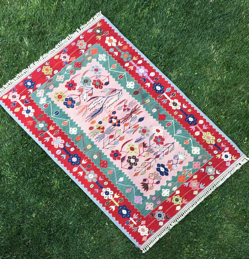 May include: A rectangular rug with a floral design. The rug features a pink central field with colorful flowers and a green border. The outer border is red with white fringe on the edges. The rug is placed on green grass.