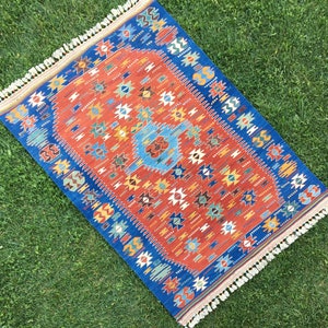 Puede incluir: Una alfombra roja, azul y blanca con un dise&ntilde;o geom&eacute;trico. La alfombra tiene un borde de flecos.