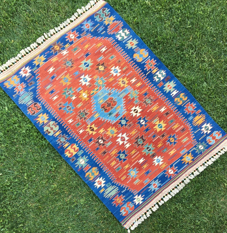 Puede incluir: Una alfombra roja, azul y blanca con un dise&ntilde;o geom&eacute;trico. La alfombra tiene un borde de flecos.