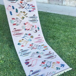 May include: A long, rectangular rug with a white background and a colorful geometric pattern. The pattern features stylized leaves and flowers in shades of pink, blue, green, red, and yellow.