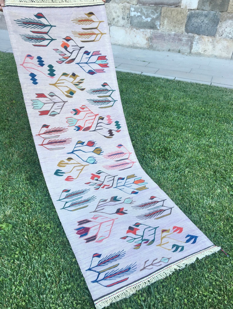 May include: A long, rectangular rug with a white background and a colorful geometric pattern. The pattern features stylized leaves and flowers in shades of pink, blue, green, red, and yellow.
