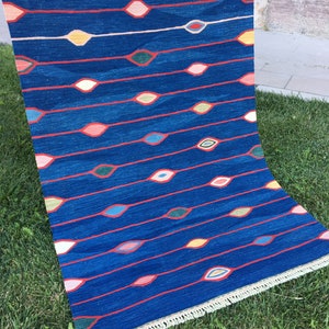 Puede incluir: Una alfombra azul con un patr&oacute;n repetitivo de dise&ntilde;os coloridos en forma de ojo. La alfombra tiene un borde de flecos.