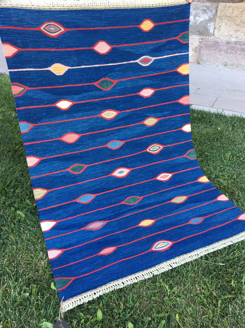 Puede incluir: Una alfombra azul con un patr&oacute;n repetitivo de dise&ntilde;os coloridos en forma de ojo. La alfombra tiene un borde de flecos.