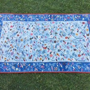 May include: A rectangular rug with a light blue central field, adorned with floral and vine patterns in various colors. The border is a darker blue with a repeating floral design and a red trim. The rug has fringed edges.