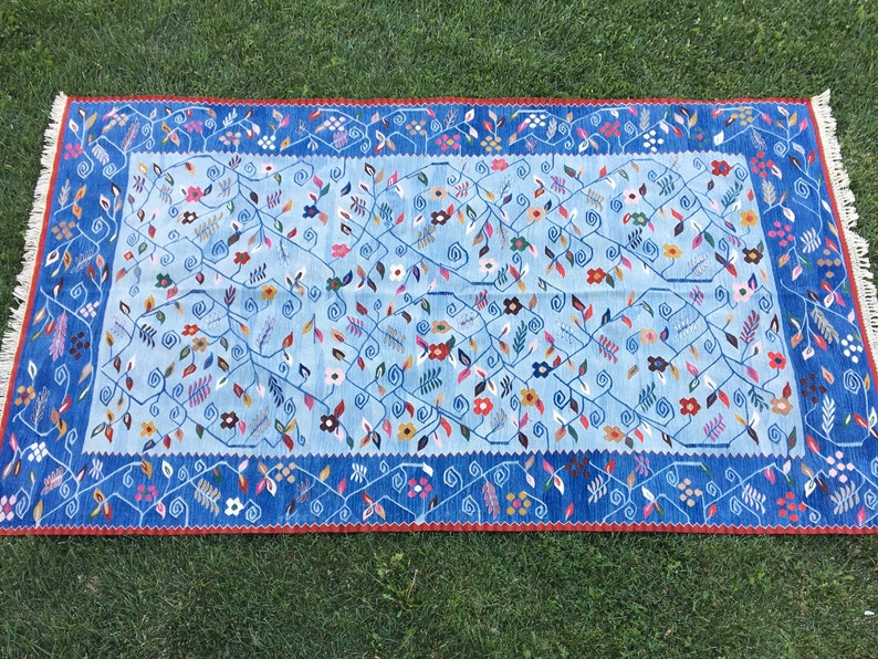 May include: A rectangular rug with a light blue central field, adorned with floral and vine patterns in various colors. The border is a darker blue with a repeating floral design and a red trim. The rug has fringed edges.