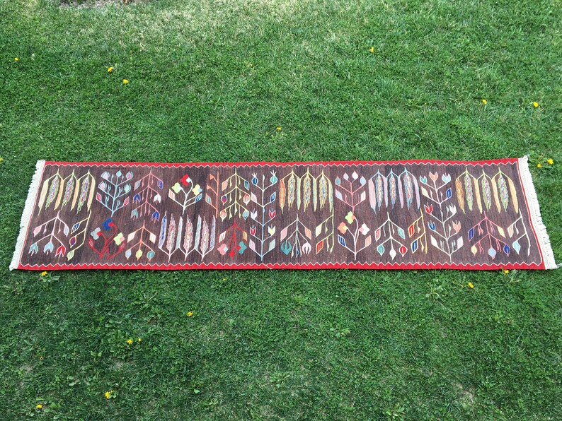 Puede incluir: Una alfombra larga y estrecha con fondo marr&oacute;n y un estampado floral colorido. La alfombra tiene un borde rojo y flecos en los extremos.