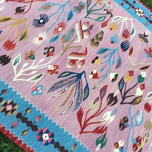 Op de afbeelding: Een roze en blauw patroon tapijt met een bloemenontwerp. Het tapijt heeft een rand van blauwe en bruine geometrische vormen.