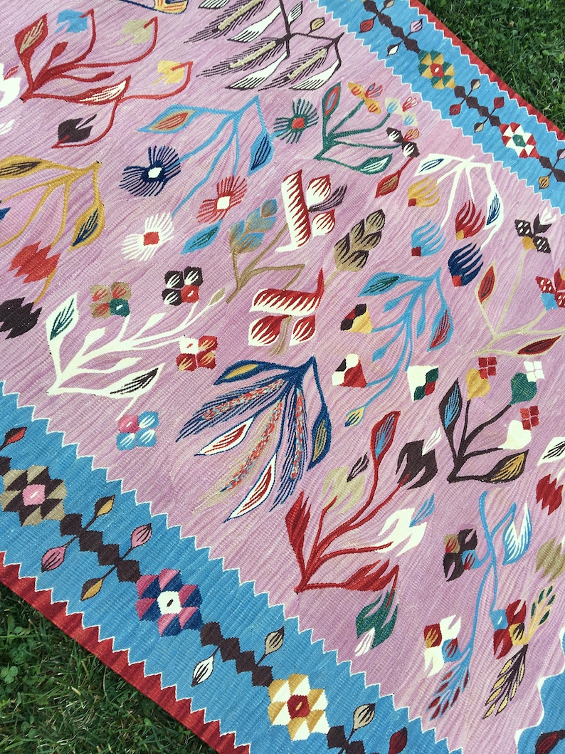 Op de afbeelding: Een roze en blauw patroon tapijt met een bloemenontwerp. Het tapijt heeft een rand van blauwe en bruine geometrische vormen.