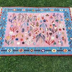 Op de afbeelding: Een roze en blauw tapijt met een bloemenpatroon. Het tapijt heeft een franje rand en is gemaakt van wol.