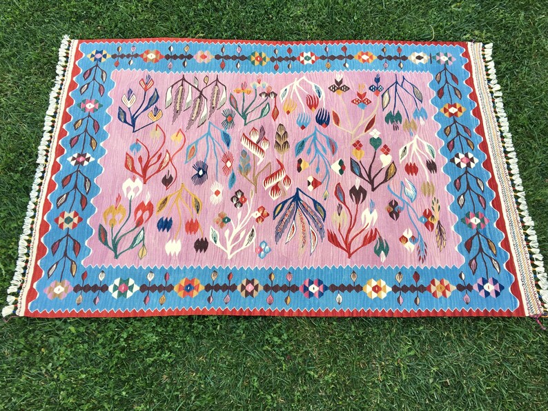 Op de afbeelding: Een roze en blauw tapijt met een bloemenpatroon. Het tapijt heeft een franje rand en is gemaakt van wol.