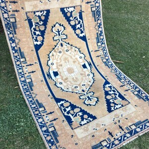 May include: A vintage rug with a beige background and a blue and white geometric pattern. The rug has a fringe border.