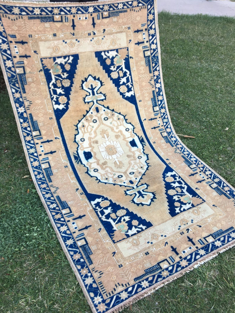 May include: A vintage rug with a beige background and a blue and white geometric pattern. The rug has a fringe border.