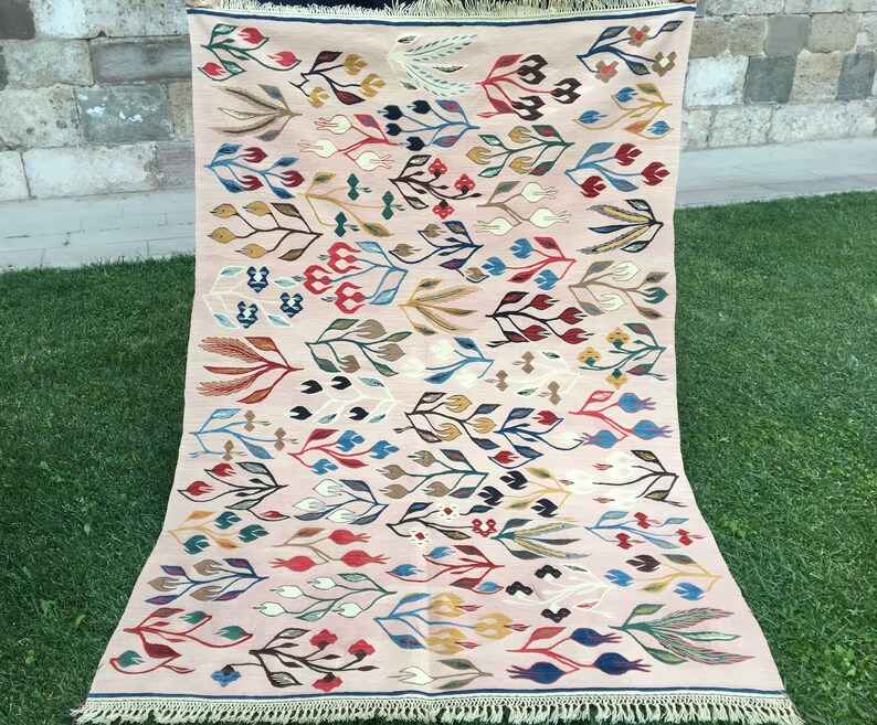 Puede incluir: Una alfombra rosa y crema con un patr&oacute;n floral. La alfombra presenta una variedad de colores, incluyendo rojo, azul, verde y amarillo. La alfombra tiene un borde de flecos.