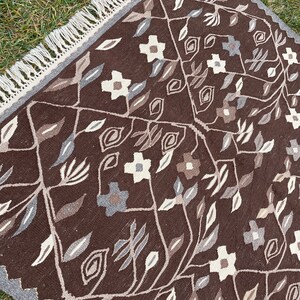 Puede incluir: Una alfombra de color marr&oacute;n y blanco con un borde de flecos. La alfombra presenta un dise&ntilde;o floral con hojas y flores.