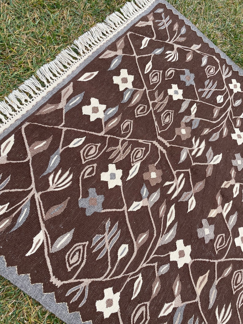 Puede incluir: Una alfombra de color marr&oacute;n y blanco con un borde de flecos. La alfombra presenta un dise&ntilde;o floral con hojas y flores.