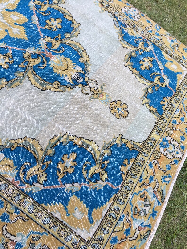 May include: A vintage rug with a distressed appearance. The rug features a central design in shades of blue and gold, with intricate floral patterns. The border is a darker blue and gold, with a cream-colored background.