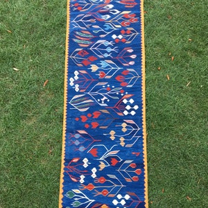 May include: A blue and yellow runner rug with a geometric pattern of stylized flowers and leaves. The rug has a fringe border.