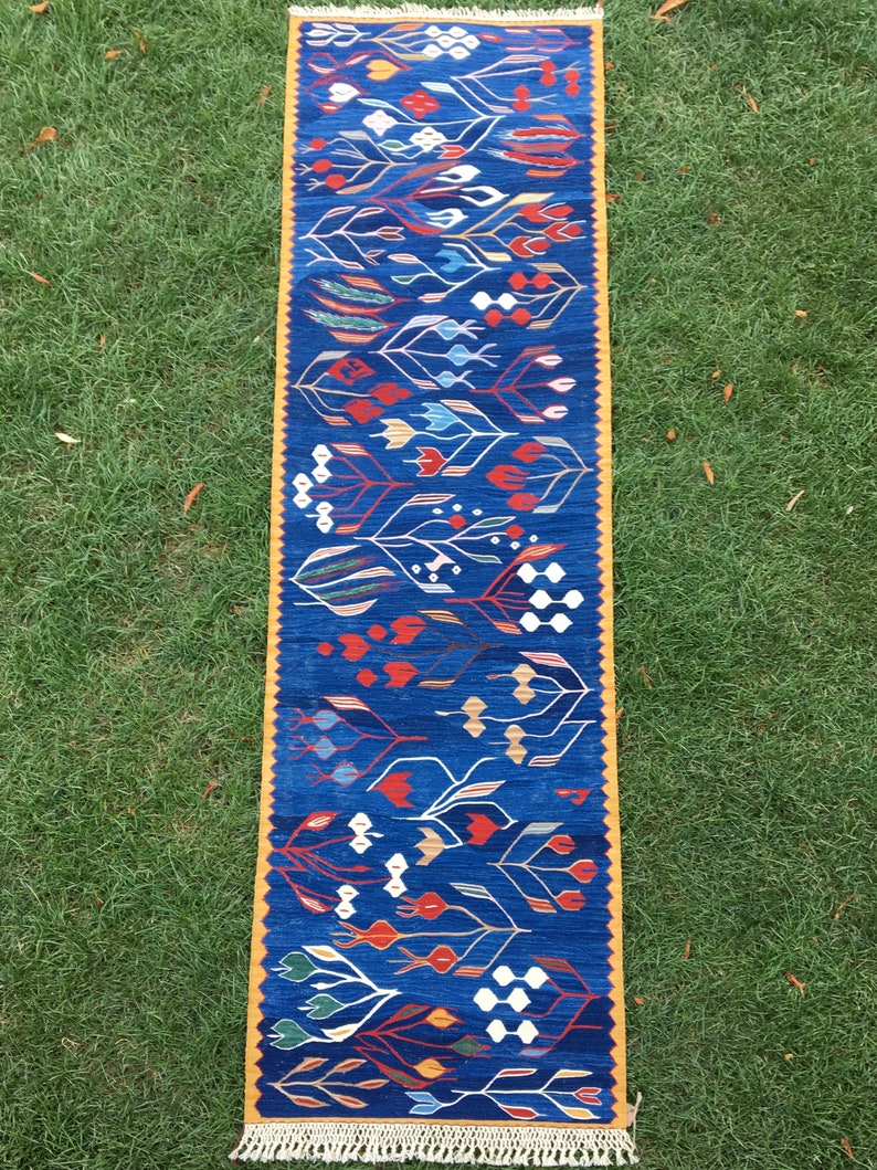 May include: A blue and yellow runner rug with a geometric pattern of stylized flowers and leaves. The rug has a fringe border.