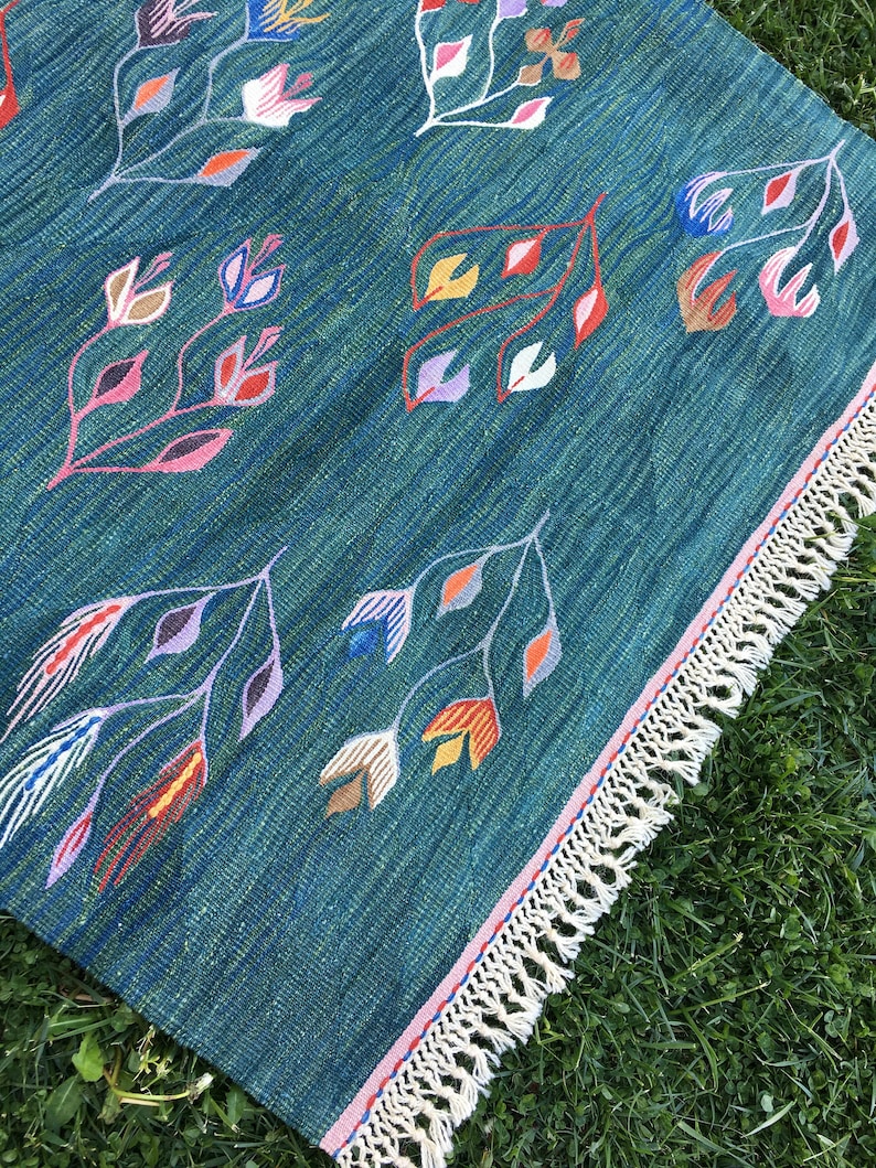 Puede incluir: Una alfombra azul verdosa con borde de flecos. La alfombra tiene un patr&oacute;n repetitivo de flores estilizadas en tonos de rosa, rojo, amarillo, naranja y morado.