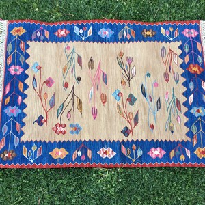 May include: A beige rug with a blue border and a floral pattern in shades of pink, red, blue, and green. The rug has fringe on all sides.