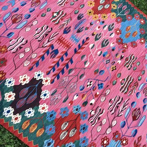 May include: A pink rug with a colourful floral pattern. The rug features a variety of colours, including red, blue, green, yellow, and white. The pattern is intricate and detailed, and the rug is made of a soft, durable material.