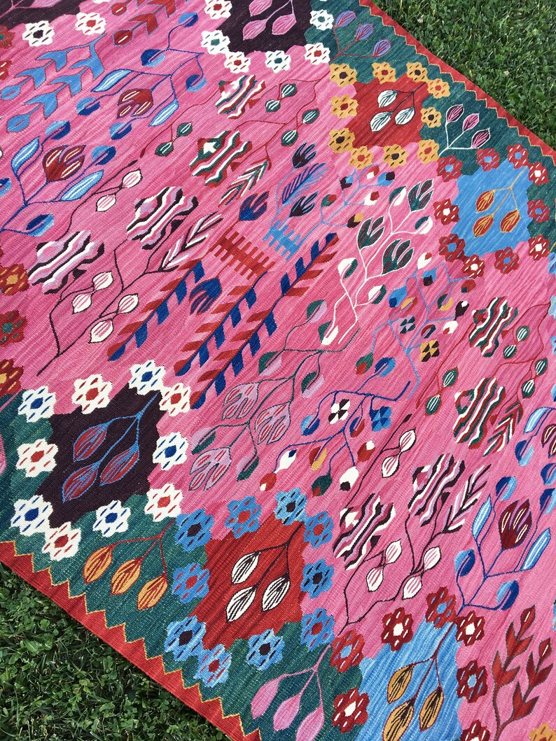 May include: A pink rug with a colourful floral pattern. The rug features a variety of colours, including red, blue, green, yellow, and white. The pattern is intricate and detailed, and the rug is made of a soft, durable material.