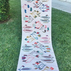 May include: A long, narrow rug with a white background and a colorful floral pattern. The rug has a fringe border and is made of wool.