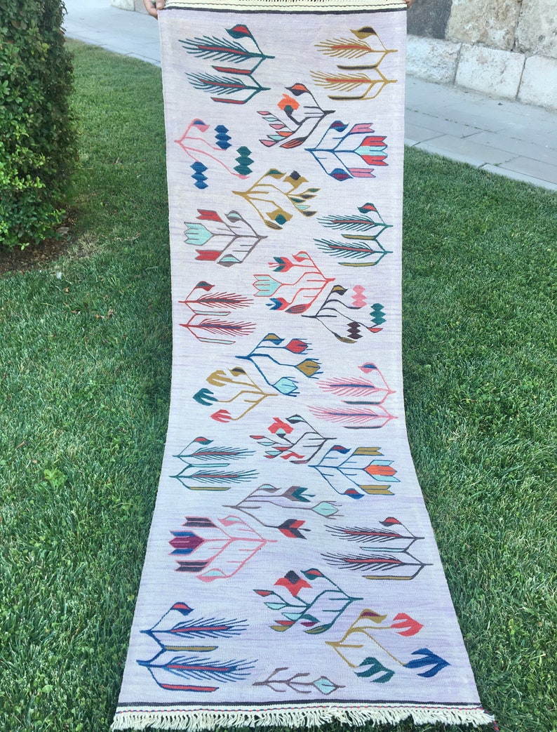 May include: A long, narrow rug with a white background and a colorful floral pattern. The rug has a fringe border and is made of wool.