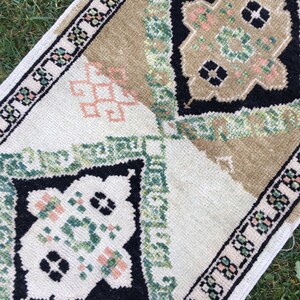May include: A small, rectangular rug with a white background and a geometric pattern in shades of green, black, and pink. The rug has a border of small, repeating patterns.
