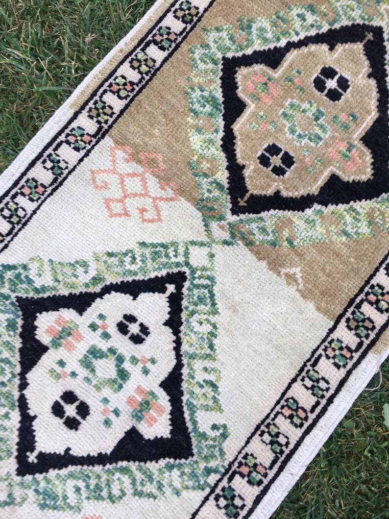 May include: A small, rectangular rug with a white background and a geometric pattern in shades of green, black, and pink. The rug has a border of small, repeating patterns.