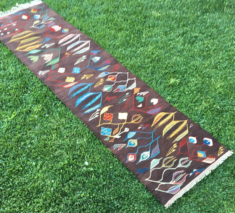 Puede incluir: Una alfombra larga y estrecha con fondo marr&oacute;n y un patr&oacute;n geom&eacute;trico colorido. La alfombra tiene flecos en ambos extremos.