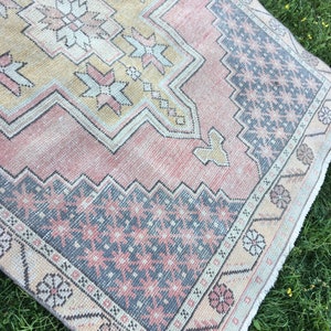 May include: A vintage-style rug with a distressed appearance. The rug features a central design in shades of peach, beige, and gray, with geometric patterns and floral accents. The border has a floral pattern. The rug is placed on a green surface.
