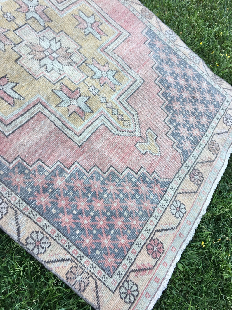 May include: A vintage-style rug with a distressed appearance. The rug features a central design in shades of peach, beige, and gray, with geometric patterns and floral accents. The border has a floral pattern. The rug is placed on a green surface.