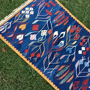 May include: A blue and yellow rug with a geometric pattern of stylized flowers and leaves. The rug has a fringe border.
